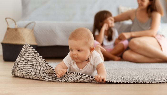 charming-baby-girl-studying-world-around-touching-carpet-while-lying-floor-living-room-adorable-kid-wearing-white-clothing-mother-sister-posing-background-web